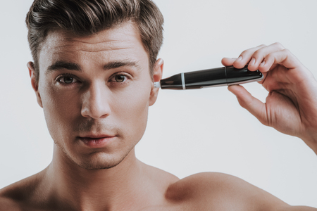 Calm handsome young Caucasian man standing shirtless against the grey wall and carefully using his nose and ear hair trimmer in left earの写真素材