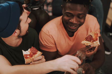 Close up portrait of caucasian bearded man and smiling afro american guy holding pizza while clinking bottles of beerの写真素材