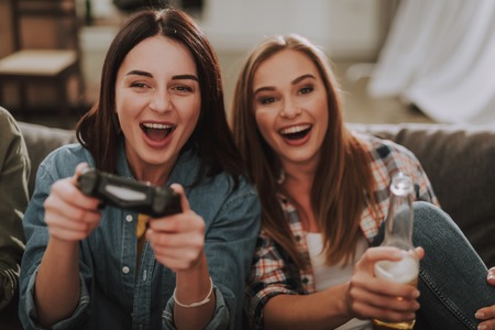 Portrait of excited young lady using joystick while her friend holding bottle of beer and laughingの写真素材