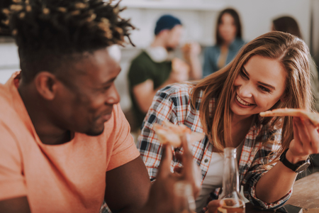 Portrait of beautiful girl holding slice of pizza and bottle of beer while looking at friend and smiling. Focus on womanの写真素材