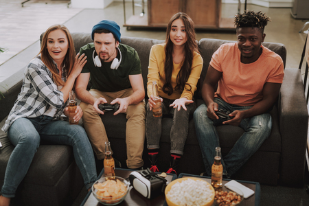 Portrait of young men using joysticks while ladies holding bottles of beer and smiling. Friends sitting on couch at homeの写真素材