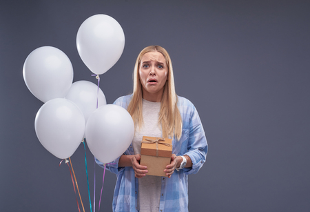 Waist up portrait of upset blond girl holding lovely gift box while standing beside white balloons.の写真素材