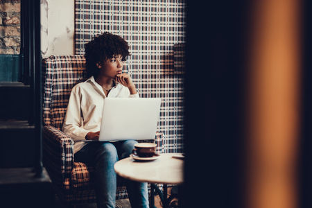Peaceful attractive woman with her calm face looking aside and sitting in comfort chair with laptopの写真素材