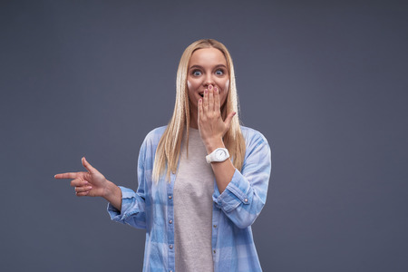Waist up portrait of charming blond girl gesturing with index finger and showing away while looking at camera with amused expression.の写真素材