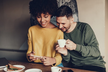 Two friends sitting in small cafe. They drinking coffee and looking at smartphone screen while laughingの写真素材