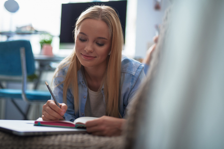 Attractive blond girl making notes while lying on couch with laptop. She smilingの写真素材
