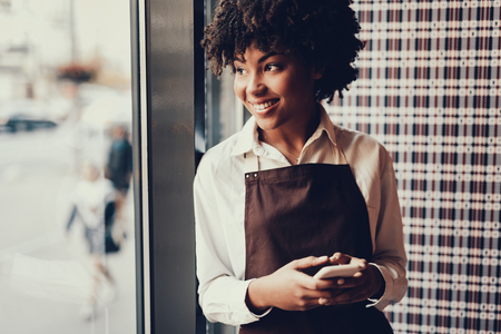 Cheerful brunette woman with cellular in hands looking through window and smiling. She standing inside small family cafeの写真素材