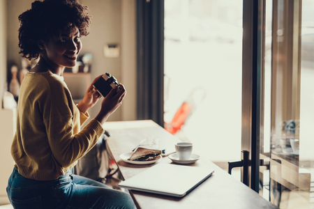 Beautiful and sweet woman sitting on cafe in yellow pullover. She holding camera in hands and smiling wideの写真素材