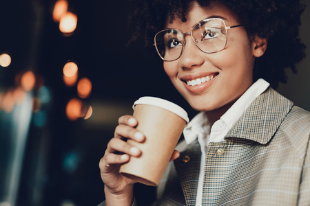 Portrait of cute woman in casual coat holding takeout beverage. She drinking coffee from brown cup of and smilingの写真素材