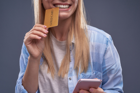 Cropped portrait of blond girl with plastic credit card between her teeth holding pink smartphone. Isolated on blue-gray backgroundの写真素材