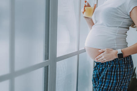 Lovely expectation. Side view of pregnant woman wearing smartwatch and holding glass of orange juice at homeの写真素材