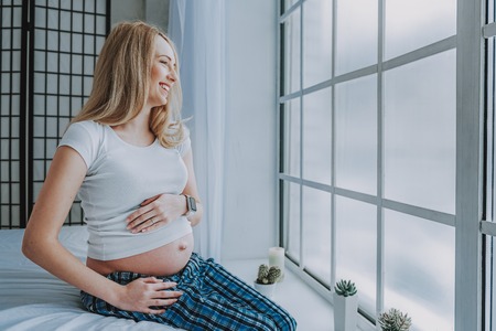Happy pregnancy. Smiling young future mother holding carefully abdomen with hands while looking out of big windowの写真素材