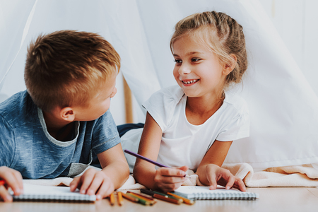 Family relationship concept. Close up portrait of smiling brother and sister looking to each other while drawing with colorful pencils on floor under self maid tentの写真素材