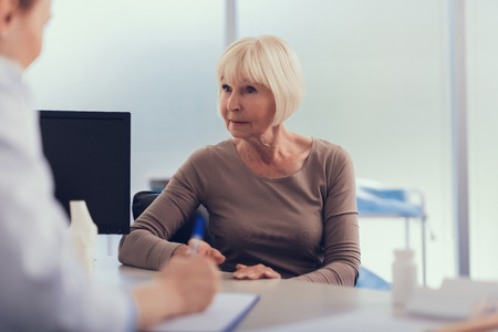 Focus on mature female listening to therapist advice during visit to clinic. She is sitting at table while female specialist is writing down her complaints into clipboardの写真素材