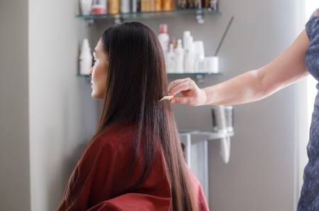 Woman is sitting in chair and wearing protective gown during treatment procedures in beauty salon. Stylist is standing behind and straightening her long hair with combの写真素材
