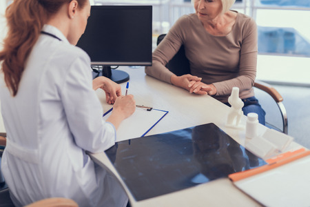 Lady and surgeon are sitting at desk while doctor is writing on tablet folder. Patient is receiving consultancy after radiography in modern office with computerの写真素材