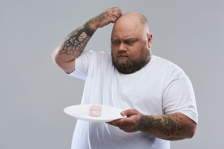 Waist up on the grey background of bearded tattooed fat man standing with a plate of marshmallow souffle and thoughtfully scratching his headの写真素材