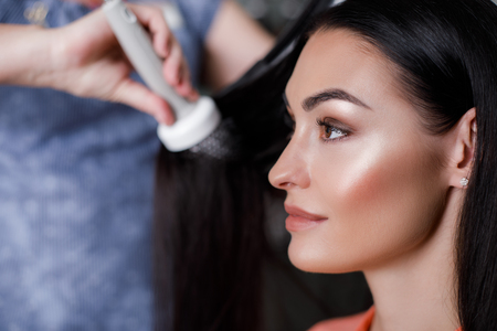 Focus on profile portrait of merry woman while doing her hair. Stylist is standing and fixing it with round brush. Copy space in left sideの写真素材