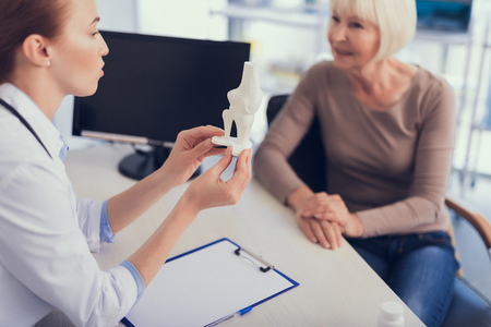 Focus on surgeon explaining leg build to patient while holding its model. They are sitting round desk with computer and documents in modern officeの写真素材