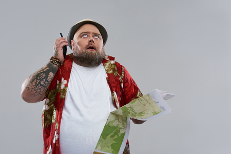 Unbearable phone talk. Tired fat bearded traveler in Hawaiian shirt looking at the sky and putting smartphone from his ear while standing isolated on the grey background with map in his handの写真素材
