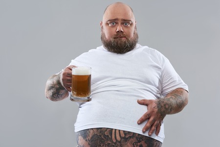 Fat middle aged bearded man putting hand on the belly while standing isolated on the grey background with a glass of beerの写真素材