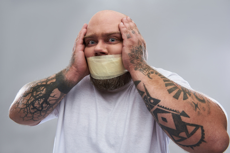 Having problems. Close up image of tattooed fat man having masking tape on his mouth and feeling unwell while touching his head isolated on the grey backgroundの写真素材