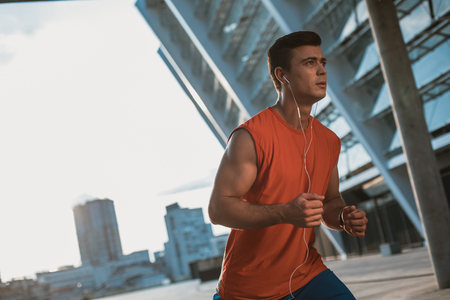 Portrait of orderly young male athlete listening music with headsets while running in city with high buildingsの写真素材