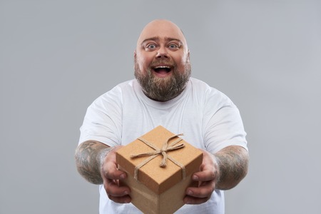 Cheerful friendly fat man with tattooed arms standing isolated on the grey background and smiling happily while giving a present in gift boxの写真素材