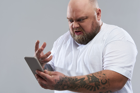 Fat bearded bold man in white T-shirt standing isolated on the grey background and gesturing while holding modern smartphone and expressing angerの写真素材