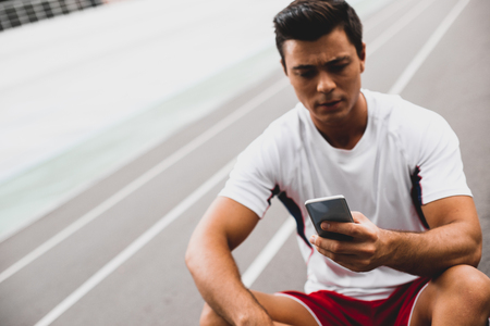Portrait of concentrated man using contemporary mobile while locating indoor in sport clothes