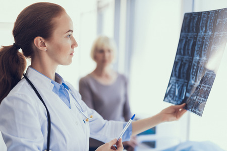 Focus on profile portrait of concentrated doctor holding radiography and looking at it. Woman is standing near and waiting for results. Copy space in right sideの写真素材