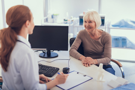 Focus on cheerful lady sitting at table while female physician is staying at table opposite to her. Medical specialist is using computer and table folderの写真素材