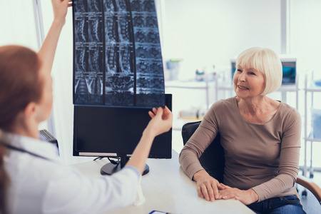 Focus on cheerful aged lady is sitting at doctor desk in medical center. Female physician is holding radiography and explaining it to patientの写真素材