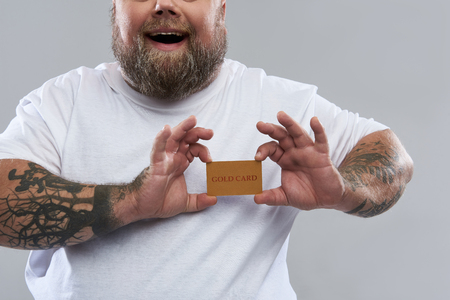 Close up of happy bearded man with tattooed arms standing alone and smiling while holding a gold card in front of his heartの写真素材