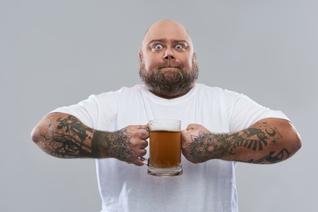 Fat bearded confident man standing in white T-shirt and opening his eyes wide while holding a big...