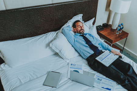 Man in shirt and tie sleeping on the hotel bed while having charts and documents next to him. Copy...