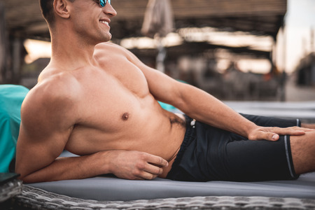 Happy relaxed young man lying shirtless on the lounge sun chair and smiling while putting one hand on the leg and looking into the distanceの写真素材