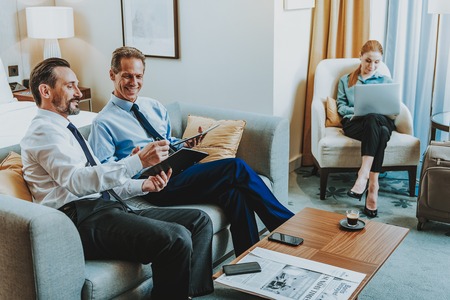 Meeting at hotel. Cheerful friendly middle aged businessmen in elegant clothes sitting on the sofa in hotel room and smiling while talking and young lady sitting with laptop on the backgroundの写真素材