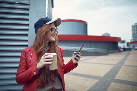 Waist up portrait of charming red-haired girl in glasses holding cup of coffee while reading messages on smartphone and smilingの写真素材