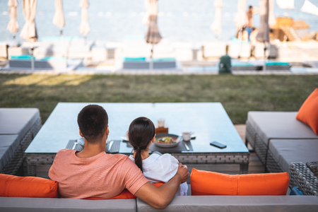 Young man sitting on terrace of outdoors cafe and putting one hand on the shoulder of his girlfriend while looking at the seaの写真素材