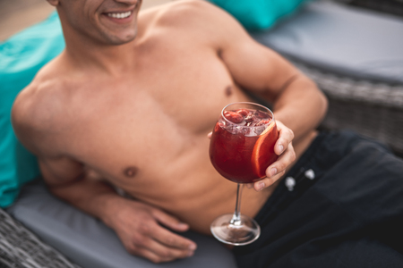 Shirtless smiling man lying on the lounge sun chair and holding a glass of tasty cold cocktail in one handの写真素材