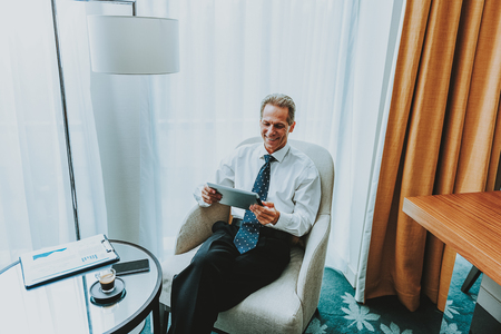 Businessman with tablet. Happy enthusiastic businessman looking glad while sitting in the armchair with coffee table in front of him and using a modern tabletの写真素材