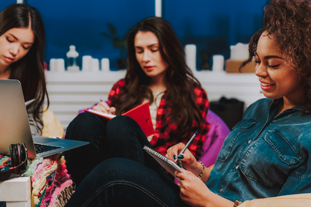 Focus on outgoing woman writing in notebook. She leaning on chair in room. Her friends sitting on backgroundの写真素材
