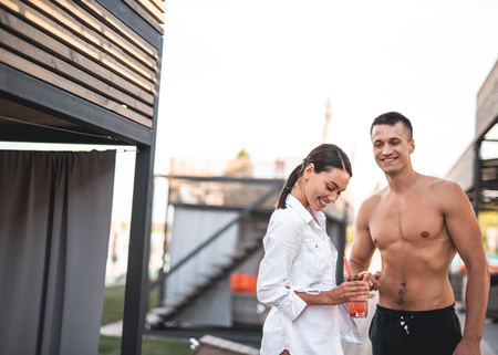 Cheerful young lady holding cocktail and laughing while her handsome shirtless boyfriend standing next to her and smiling. Copy space on the left sideの写真素材