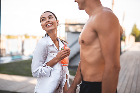 Happy young woman smiling and looking away while holding the hand of her handsome shirtless boyfriend and enjoying tasty cocktail outdoorsの写真素材