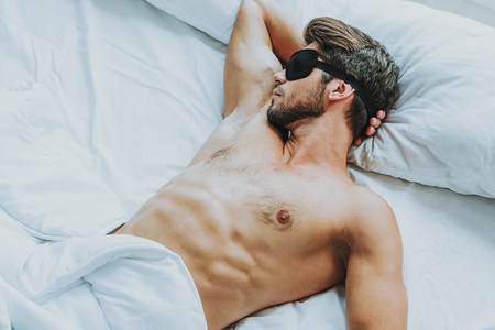 Relaxation and comfort in the morning. Close up portrait of young male with naked muscular torso sleeping in bed with eyes covering by blindfoldの写真素材