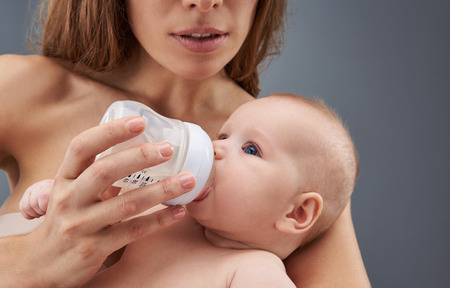 Cropped and close up photo of feeding baby from plastic bottle. Mama standing isolated on gray background and holding newborn child on handsの写真素材