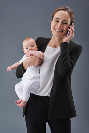 Vertical photo of mommy in formalwear standing isolated on gray background, speaking on telephone and holding small child on handの写真素材