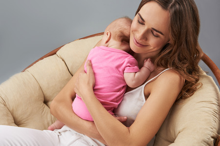 Attractive mom wearing in white everyday clothes sitting in armchair isolated on gray background. She closing eyes and holding her small child in handsの写真素材
