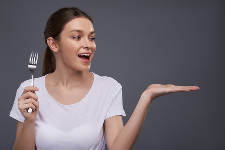 Portrait of beautiful young lady with eating utensil looking away with cheerful expression and keeping mouth open. Isolated on gray backgroundの写真素材